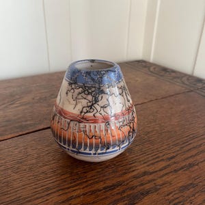 May include: A small, handcrafted ceramic vase with a unique design. The vase features a blue rim and base, with orange and white bands in the middle. Black, lightning-like patterns cover the vase's surface, adding a striking contrast. The vase sits on a wooden table.