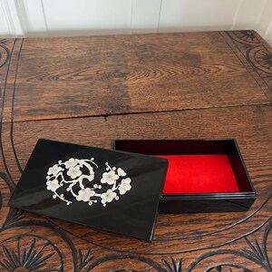 May include: A black lacquered box with a silver floral design on the lid. The interior is lined with red fabric. The box is open, revealing the interior. The box is on a wooden surface.