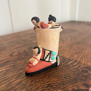 May include: A handcrafted ceramic boot figurine with three small figures. The boot is painted in shades of red, black, and tan, with turquoise accents. The figures are depicted in traditional attire.