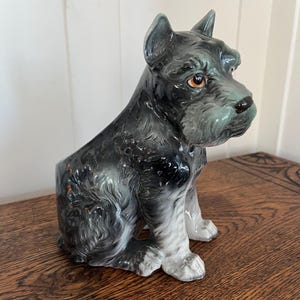 May include: A ceramic figurine of a Schnauzer dog in a seated position. The dog has a black and gray coat with detailed fur texture, brown eyes, and a dark nose. The figurine is on a wooden surface.