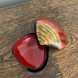 May include: A small, open, square-shaped box with a red interior and a black rim. The lid is a rounded shape with a red and gold design. The box is on a wooden surface.