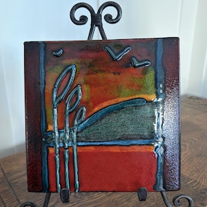 May include: A square ceramic tile featuring a glossy, multi-colored landscape design with raised blue lines depicting tall reeds and birds in flight. The tile is displayed on a black metal stand with decorative scrollwork at the top and base.