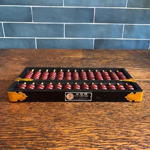 May include: A vintage abacus with a black frame and yellow corner accents. The abacus features rows of red beads, and a small label with Chinese characters. The abacus is a traditional calculating tool.