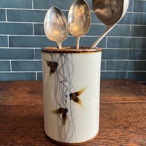 May include: A ceramic utensil holder with a cream-colored base, brown trim, and a painted design of brown and blue abstract shapes. Three silver spoons and a ladle are inside the holder, which is sitting on a wooden surface.