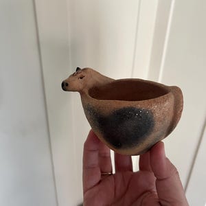 May include: A handmade, brown ceramic bowl in the shape of a cow. The bowl has a dark brown patch on the side and a small, raised head with horns. The interior is hollow, and the bowl is held in a hand.