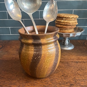 May include: A ceramic utensil holder with brown and yellow stripes holds three silver spoons. A stack of golden-brown cookies sits on a decorative silver cake stand, suggesting a kitchen or dining setting.