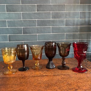 May include: A collection of six vintage wine glasses in various shades of amber, brown, and red. Each glass features a unique design, including textured patterns and intricate stem details. The glasses are arranged on a wooden surface.