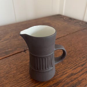 May include: A small, dark brown ceramic creamer with a white interior. The creamer has a handle and a decorative band of vertical lines around the middle. It sits on a wooden surface.