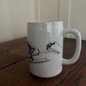 Puede incluir: Taza de cerámica blanca moteada con asa curva. La taza presenta una ilustración en blanco y negro de dos esquiadores en acción. La taza está sobre una superficie de madera.