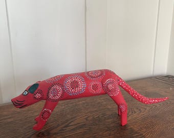 May include: A red wooden sculpture of a quadruped animal, possibly a dog or a cat, with a long tail. The body is decorated with white, blue, and purple circular patterns. The sculpture is on a wooden surface.