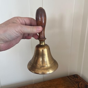 May include: A vintage brass bell with a wooden handle. The bell has a classic shape with a flared base and a dark brown, polished wooden handle. The brass has a warm, aged patina. The bell is held by a hand.