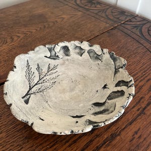 May include: A handmade, off-white ceramic bowl with a textured surface and a scalloped edge. The bowl features a black botanical design and dark gray accents. The bowl is displayed on a wooden table.