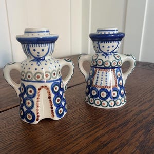 May include: Two ceramic figurines in a folk art style. Each figure has a blue hat, arms, and a patterned body with blue, brown, and green accents. The figurines are on a wooden surface.