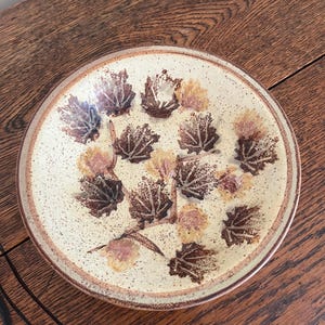 May include: A round ceramic plate with a beige speckled background and a brown rim. The plate is decorated with brown and tan leaf designs. The plate is sitting on a wooden surface.