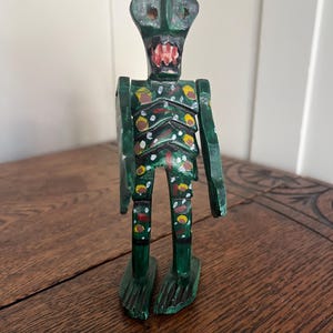 May include: A green wooden figurine with a humanoid form. The figure has a rectangular head with two eye holes and a red mouth. The body is painted with yellow, white, and black accents. The feet are carved with a textured design.