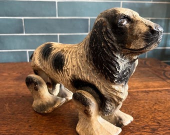 May include: A vintage dog figurine with a spotted coat in black and tan. The dog is posed with its front legs extended, and the figurine has a distressed finish. The dog is sitting on a wooden surface.