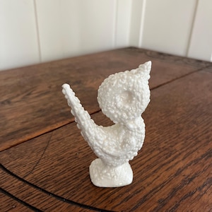 May include: A white ceramic bird figurine with a textured, bubbled surface. The bird has a curved body, a pointed beak, and a small base. The figurine is displayed on a wooden surface.