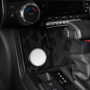 May include: A black textured rubber mat with a geometric pattern installed in a vehicle center console storage area, featuring a circular white magnetic charging disc mounted in the center, positioned near the gear shifter and climate controls.