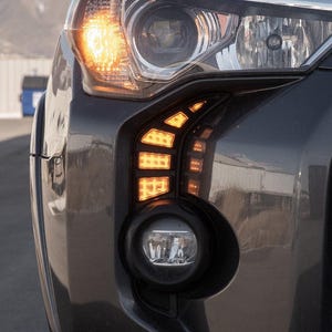 May include: Close-up of a dark grey vehicle's front end, highlighting the headlight and fog light. The headlight is illuminated with a bright orange light, and the fog light is a round shape. The vehicle's body has a glossy finish.