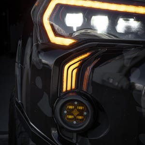 May include: Close-up of a black vehicle's headlight assembly. The headlight features bright white and amber LED lights. A round, amber-coloured fog light is mounted below the headlight. The vehicle has a camouflage pattern.