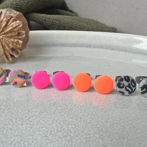 Könnte beinhalten: Ein Set mit acht bunten und gemusterten Ohrringen. Die Ohrringe sind aus Polymer Clay gefertigt und haben verschiedene Designs, darunter rosa, orange und grau mit schwarzen Mustern. Die Ohrringe sind alle rund und haben einen silbernen Stift.