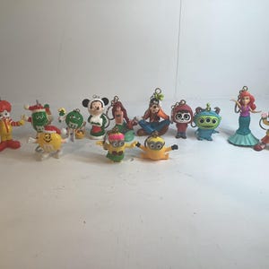 Könnte beinhalten: Eine Sammlung farbenfroher Figuren, darunter Mickey Mouse, Arielle, Minions und andere. Jede Figur ist detailliert und hat eine kleine Schlaufe zum Aufhängen. Die Figuren sind auf einer weißen Oberfläche angeordnet.
