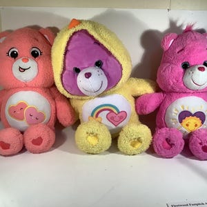 May include: Three Care Bears plush toys. The first is pink with a heart design and the text "Love You". The second is yellow with a rainbow and heart design. The third is pink with a sun and heart design.