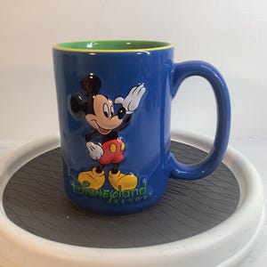 May include: A blue ceramic mug featuring a raised, three-dimensional graphic of Mickey Mouse waving. The text Disneyland Resort is printed in green below the character. The interior of the mug is a bright lime green with a yellow rim.