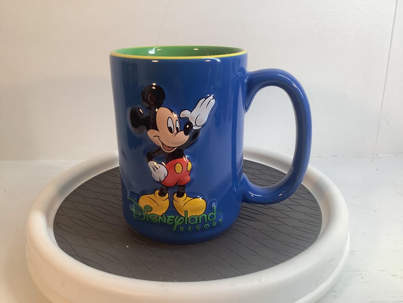 May include: A blue ceramic mug featuring a raised, three-dimensional graphic of Mickey Mouse waving. The text Disneyland Resort is printed in green below the character. The interior of the mug is a bright lime green with a yellow rim.