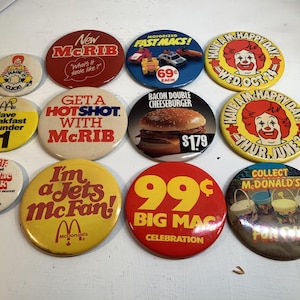 Puede incluir: Una colección de botones promocionales vintage de McDonald's. Los botones presentan varios diseños, incluyendo McRib, Big Mac y Ronald McDonald. Algunos botones tienen texto como "Desayuno por menos de 1$" y "I'm a Jets McFan!"
