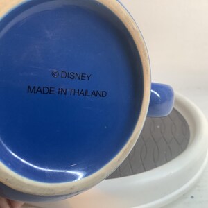 May include: The underside of a blue ceramic mug featuring a circular base with a white rim. The text printed in black on the blue surface reads &copy; DISNEY and MADE IN THAILAND. A portion of the mug handle is visible on the right side of the frame.