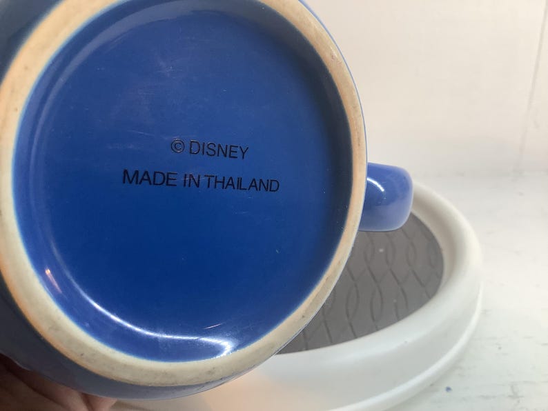 May include: The underside of a blue ceramic mug featuring a circular base with a white rim. The text printed in black on the blue surface reads &copy; DISNEY and MADE IN THAILAND. A portion of the mug handle is visible on the right side of the frame.