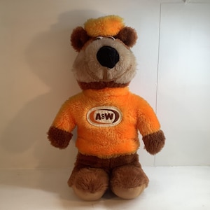 Pode incluir: Um urso de pelúcia vestindo um suéter laranja com o logotipo A&W, calças marrons e um chapéu combinando. O urso tem um rosto bege, orelhas marrons e um nariz preto. O bicho de pelúcia está em pé.