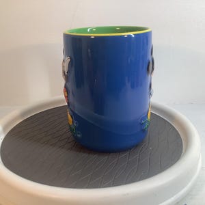 May include: A cylindrical ceramic mug with a glossy blue exterior and a bright green interior rim. The mug features raised, three-dimensional character figures molded onto the side, showcasing vibrant colors including yellow, black, and white accents.