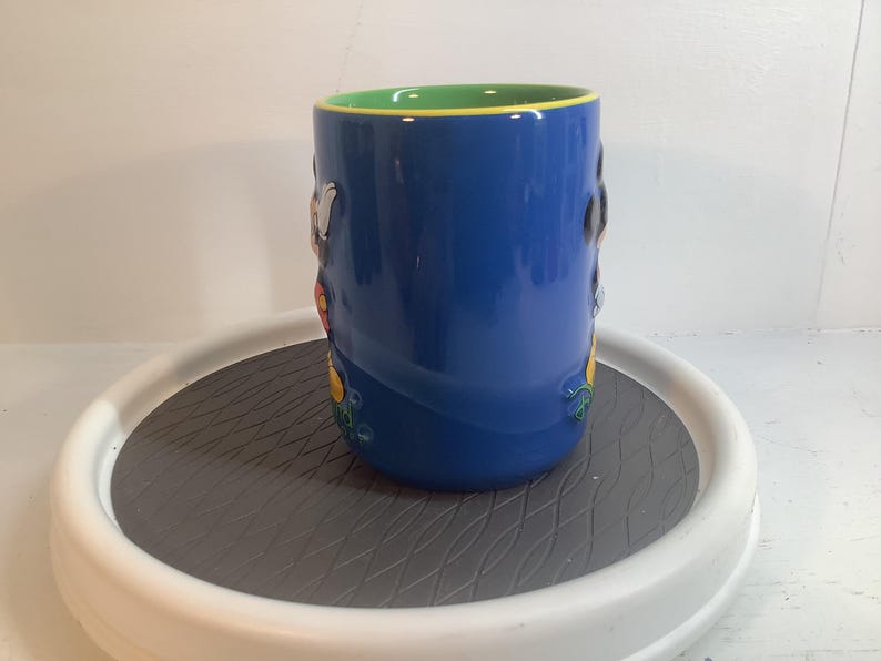 May include: A cylindrical ceramic mug with a glossy blue exterior and a bright green interior rim. The mug features raised, three-dimensional character figures molded onto the side, showcasing vibrant colors including yellow, black, and white accents.