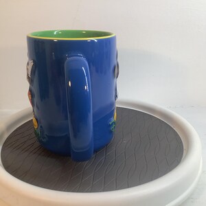 May include: A blue ceramic mug featuring a bright green interior and a large, curved handle. The exterior surface displays raised, colorful character figures in relief, including a white and black figure and a yellow element, set against the blue.