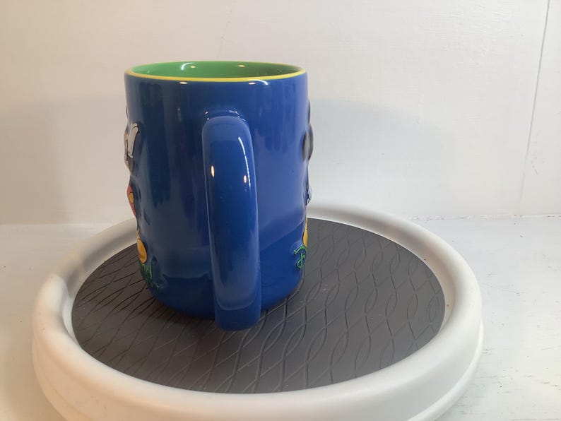 May include: A blue ceramic mug featuring a bright green interior and a large, curved handle. The exterior surface displays raised, colorful character figures in relief, including a white and black figure and a yellow element, set against the blue.