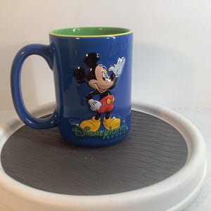 May include: A blue ceramic mug featuring a raised, three-dimensional figure of Mickey Mouse waving. The mug has a bright green interior and the text Disneyland Resort printed in green script at the base of the character on the front side.