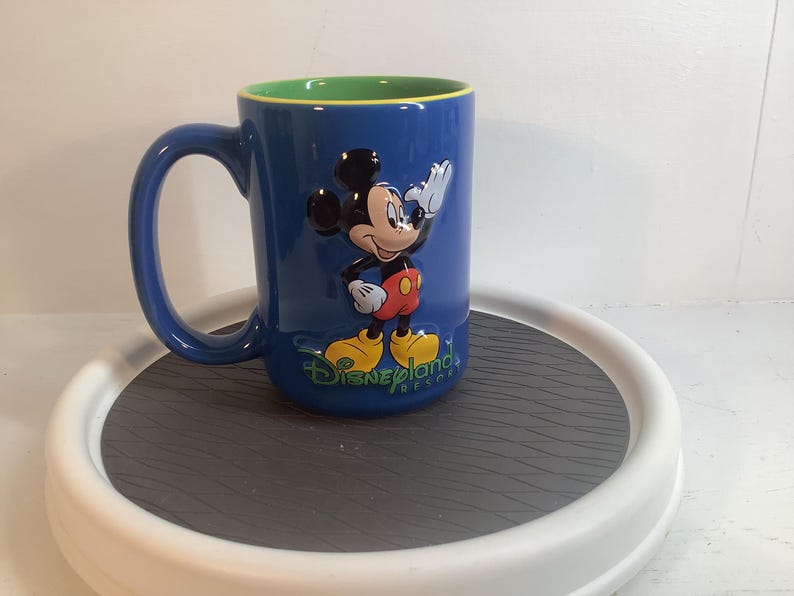 May include: A blue ceramic mug featuring a raised, three-dimensional figure of Mickey Mouse waving. The mug has a bright green interior and the text Disneyland Resort printed in green script at the base of the character on the front side.