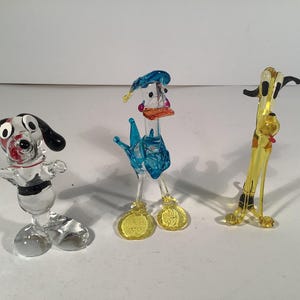 May include: Three glass figurines of cartoon characters. A blue bird with a yellow beak and feet, a white dog with black spots and ears, and a yellow dog with black ears and a red collar.