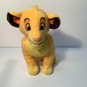 Puede incluir: Un juguete de peluche de Simba, el cachorro de león de El Rey León de Disney. Simba está sentado con las patas metidas delante de él. Tiene un pelaje amarillo y naranja, orejas negras con detalles naranjas y una nariz rosa.