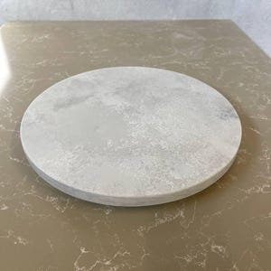 Puede incluir: Una tabla redonda de piedra gris claro con una superficie texturizada. La tabla es circular y se encuentra sobre una superficie marrón claro. La tabla es adecuada para servir queso, aperitivos u otras creaciones culinarias.