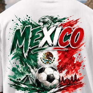 K&ouml;nnte beinhalten: Ein wei&szlig;es T-Shirt mit einer gro&szlig;en Grafik auf der R&uuml;ckseite, die das Wort MEXICO in fetten Buchstaben zeigt. Das Design umfasst einen Fu&szlig;ball, die mexikanische Flagge, einen Adler, eine Pyramide und eine Skyline im Farbklecks-Stil.