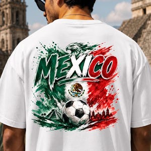 K&ouml;nnte beinhalten: Ein wei&szlig;es T-Shirt mit einer gro&szlig;en Grafik auf der R&uuml;ckseite, die das Wort MEXICO in fetten Buchstaben zeigt. Das Design umfasst einen Fu&szlig;ball, die mexikanische Flagge, einen Adler, eine Pyramide und eine Skyline im Farbklecks-Stil.