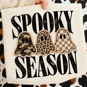 May include: A cream-colored tote bag featuring the black text SPOOKY SEASON. Three ghost-shaped patches are centered, each with black oval eyes and a thick rope border. The ghosts feature camouflage, leopard print, and tan checkerboard patterns.