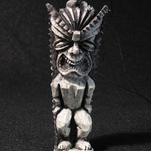 May include: A small, off-white tiki statue with a menacing expression. The carved figure features a detailed headdress, a wide, toothy grin, and stylized arms and legs. A black string is attached at the top for hanging. The statue is set against a dark background.
