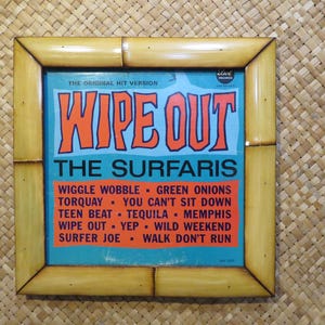 May include: A framed vintage album cover for "Wipe Out" by The Surfaris. The album art features the title in orange block letters over a blue background. The frame is made of light brown bamboo.