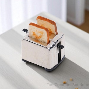 Puede incluir: Una tostadora cromada en miniatura con dos rebanadas de pan tostado. El pan es de color marrón dorado con un aspecto ligeramente quemado. La tostadora tiene palancas negras y está sobre una superficie clara. La imagen está bien iluminada.