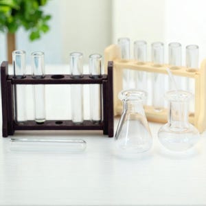 May include: A collection of miniature laboratory glassware, including test tubes in wooden racks and small flasks. The clear glass contrasts with the dark brown and light wood racks, creating a scientific aesthetic. A single test tube lies horizontally.