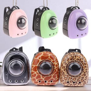Puede incluir: Una colección de mochilas transportadoras para mascotas en varios colores y estampados. Las mochilas tienen una ventana transparente en forma de burbuja. Los colores incluyen rosa, verde, morado, negro, estampado de jirafa y leopardo.